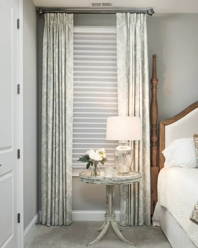 Bedroom corner with a window, patterned curtains, and a striped blind. A round table with a lamp and flowers is next to a bed with a wooden post and white bedding.